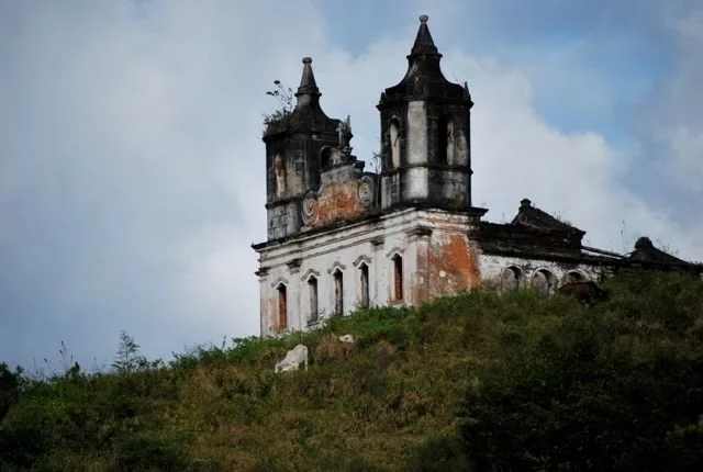 Fachada de uma igreja antiga e abandonada, parcialmente coberta por plantas, simbolizando o abandono e a passagem do tempo.